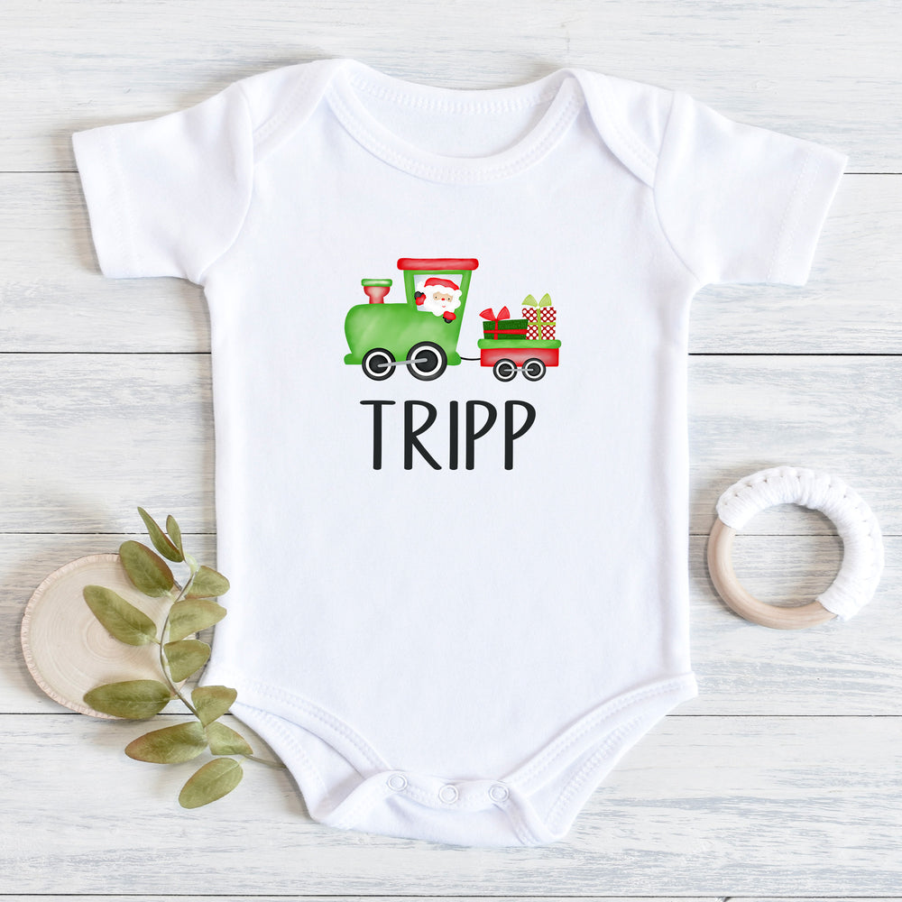 White short-sleeve baby bodysuit laid flat on a light wood surface, printed with a green Christmas train carrying Santa and gifts above the name 'TRIPP' in black letters. A small green leaf sprig and a white wooden teething ring are placed beside it.