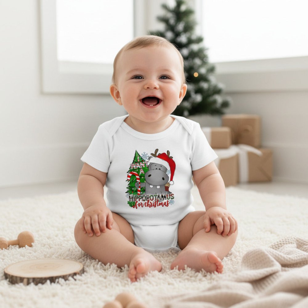 A white short-sleeve baby bodysuit laid flat on a light wood surface, featuring a Christmas hippopotamus graphic with a red Santa hat, green tree, and the text 'HIPPOPOTAMUS for Christmas.' A wooden teething ring and green leaves are placed beside it as styling props.