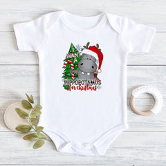 A white short-sleeve baby bodysuit laid flat on a light wood surface, featuring a Christmas hippopotamus graphic with a red Santa hat, green tree, and the text 'HIPPOPOTAMUS for Christmas.' A wooden teething ring and green leaves are placed beside it as styling props.