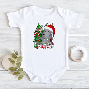 A white short-sleeve baby bodysuit laid flat on a light wood surface, featuring a Christmas hippopotamus graphic with a red Santa hat, green tree, and the text 'HIPPOPOTAMUS for Christmas.' A wooden teething ring and green leaves are placed beside it as styling props.