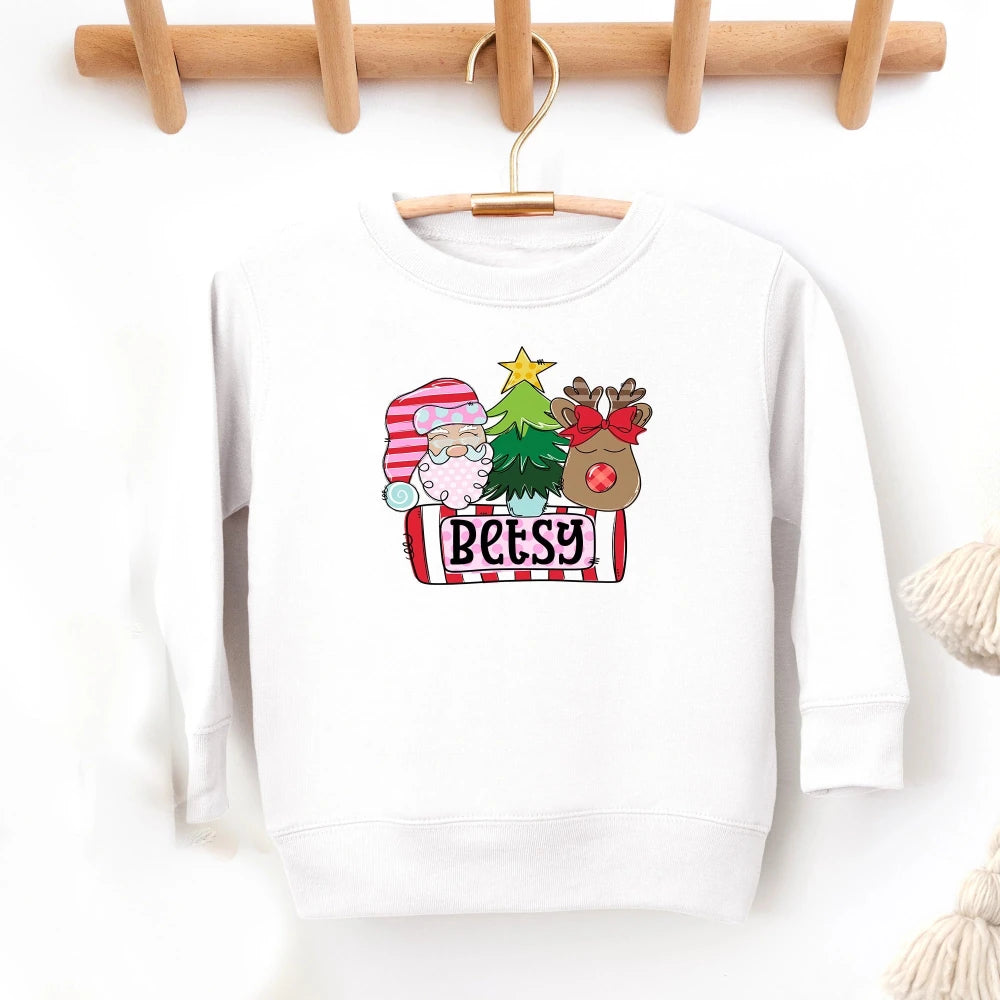 A white crewneck sweatshirt hanging on a wooden hanger against a white wall, featuring a colorful Christmas-themed graphic on the front with a sheep in a Santa hat, a Christmas tree, a reindeer, and the name 'Betsy' on a banner.