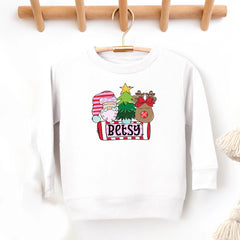 A white crewneck sweatshirt hanging on a wooden hanger against a white wall, featuring a colorful Christmas-themed graphic on the front with a sheep in a Santa hat, a Christmas tree, a reindeer, and the name 'Betsy' on a banner.