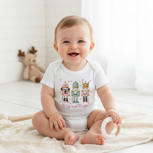 A smiling baby sitting on a cream knit blanket in a bright room, wearing a white short-sleeve bodysuit with a colorful nutcracker graphic and the words 'Merry and Bright'.
