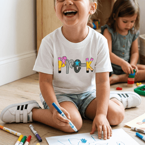 A young girl sitting on a wooden floor laughing while wearing a white graphic t-shirt with the word 'PROK' in colorful letters. She is holding a blue marker, with crayons on the floor around her and another child in the background.