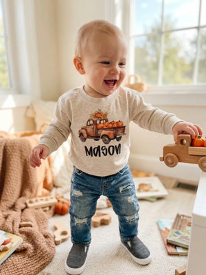 A baby standing in a bright room wearing a cream long-sleeve shirt with a pumpkin truck graphic and the name 'MASON' printed below it.
