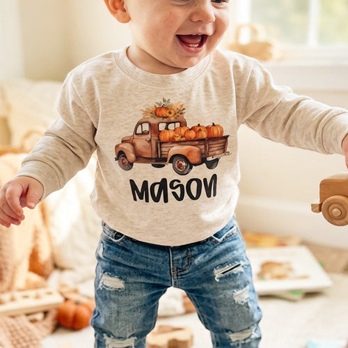A baby wearing a light beige long-sleeve shirt with a graphic of a brown pickup truck carrying pumpkins and the name 'MASON' printed below it, paired with distressed blue jeans in a cozy indoor setting.
