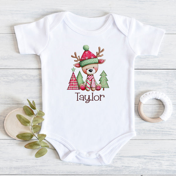 A white short-sleeve baby bodysuit laid flat on a light wood surface, printed with a cute holiday reindeer wearing a red and green hat and scarf, with the name 'Taylor' below. Decorative leaves and a wooden teether are placed beside it.