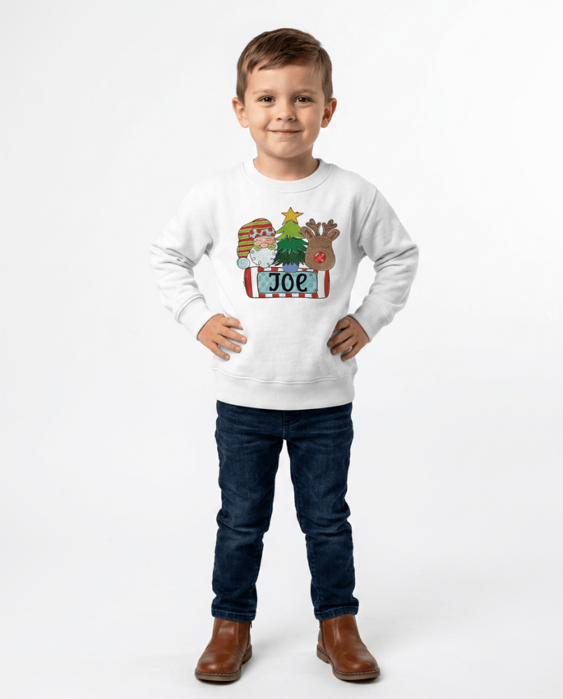 A young boy wearing a white crewneck sweatshirt with a personalized Christmas graphic with Santa, a Christmas tree, and Reindeer with a custome name printed on it.