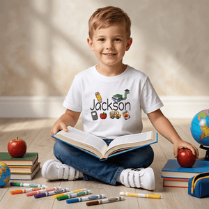 A young boy sitting cross-legged on a wooden floor surrounded by school supplies, wearing a white t-shirt with a custom graphic that reads 'Jackson' with school-themed icons. He holds an open book, with apples, books, and markers around him.