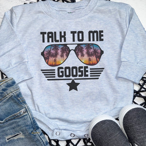 A light heather gray baby bodysuit with a graphic print reading 'TALK TO ME GOOSE' and sunglasses filled with a sunset palm tree scene, styled flat-lay with denim shorts and gray slip-on shoes.